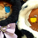 viktor plushie wearing a black cat-themed capelet with purple bows and eilhart plushie wearing a fluffy cow-themed capelet